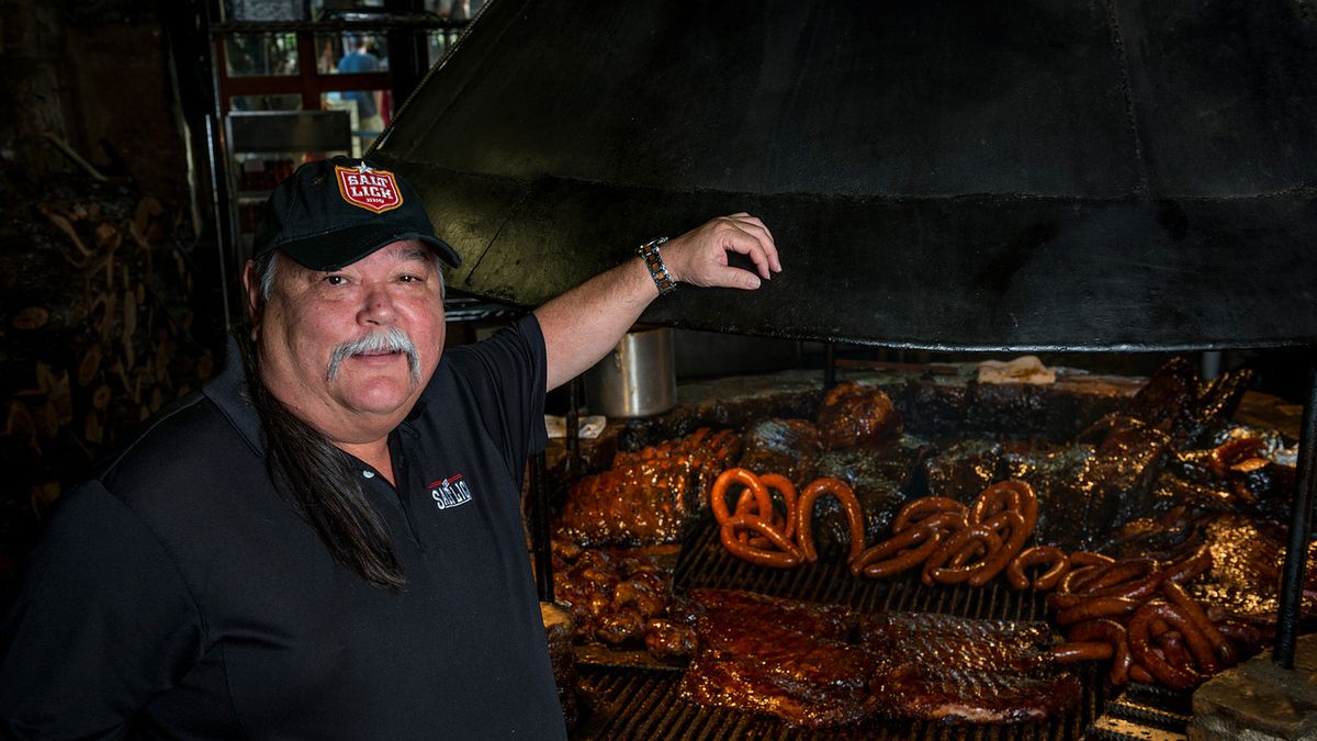 Scott Roberts, the founder of the Salt Lick BBQ, stands in front of a pit stacked with smoked meats.