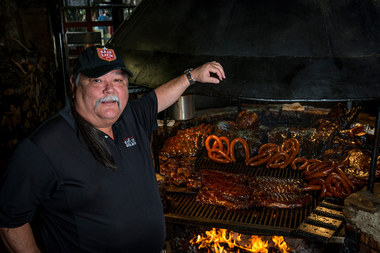 Scott Roberts, the founder of the Salt Lick BBQ, stands in front of a pit stacked with smoked meats.