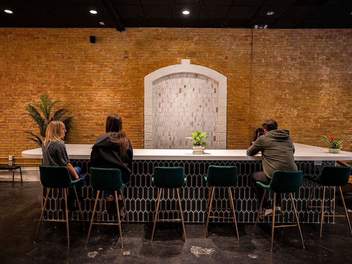 Three customers sit at a bar with a navy blue tile on the bottom and an exposed brick wall behind. 