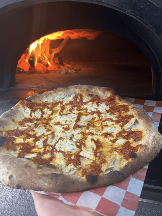 A pizza sits in front of a fire-burning oven.