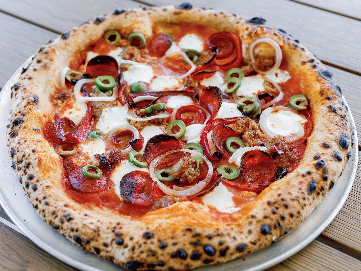 A pizza sits on a white plate. It is topped with pepperoni, olives, and onions.