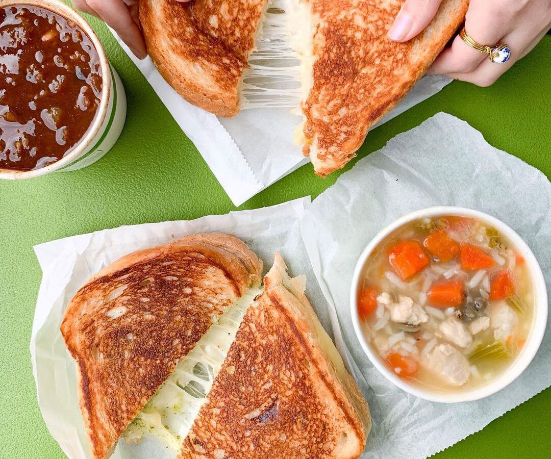 A grilled cheese that is split in two sits next to a bowl of chicken and rice soup with carrots and celery.