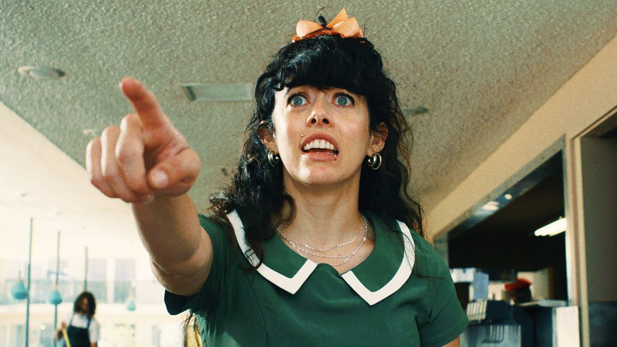 A woman in a waitress uniform stands with her mouth open, pointing towards the camera.