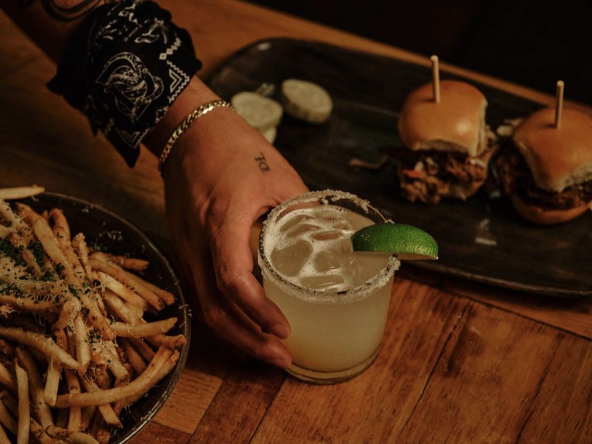 A hand sets a margarita down on a table between a plate of sliders and fries.