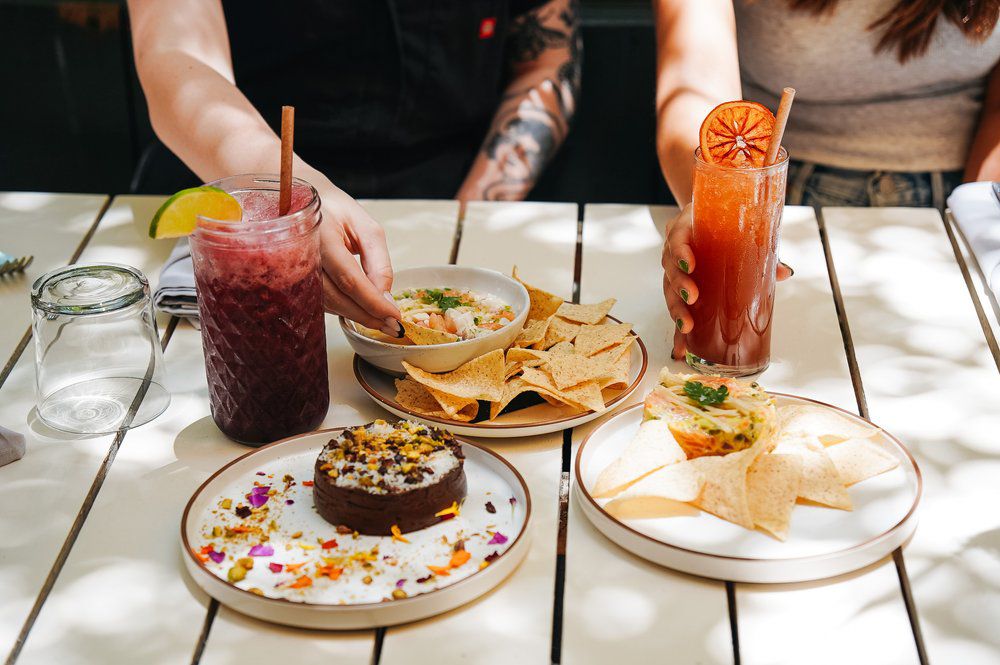 Two people sit at a white board table with food and two colorful cocktails in front of them. 