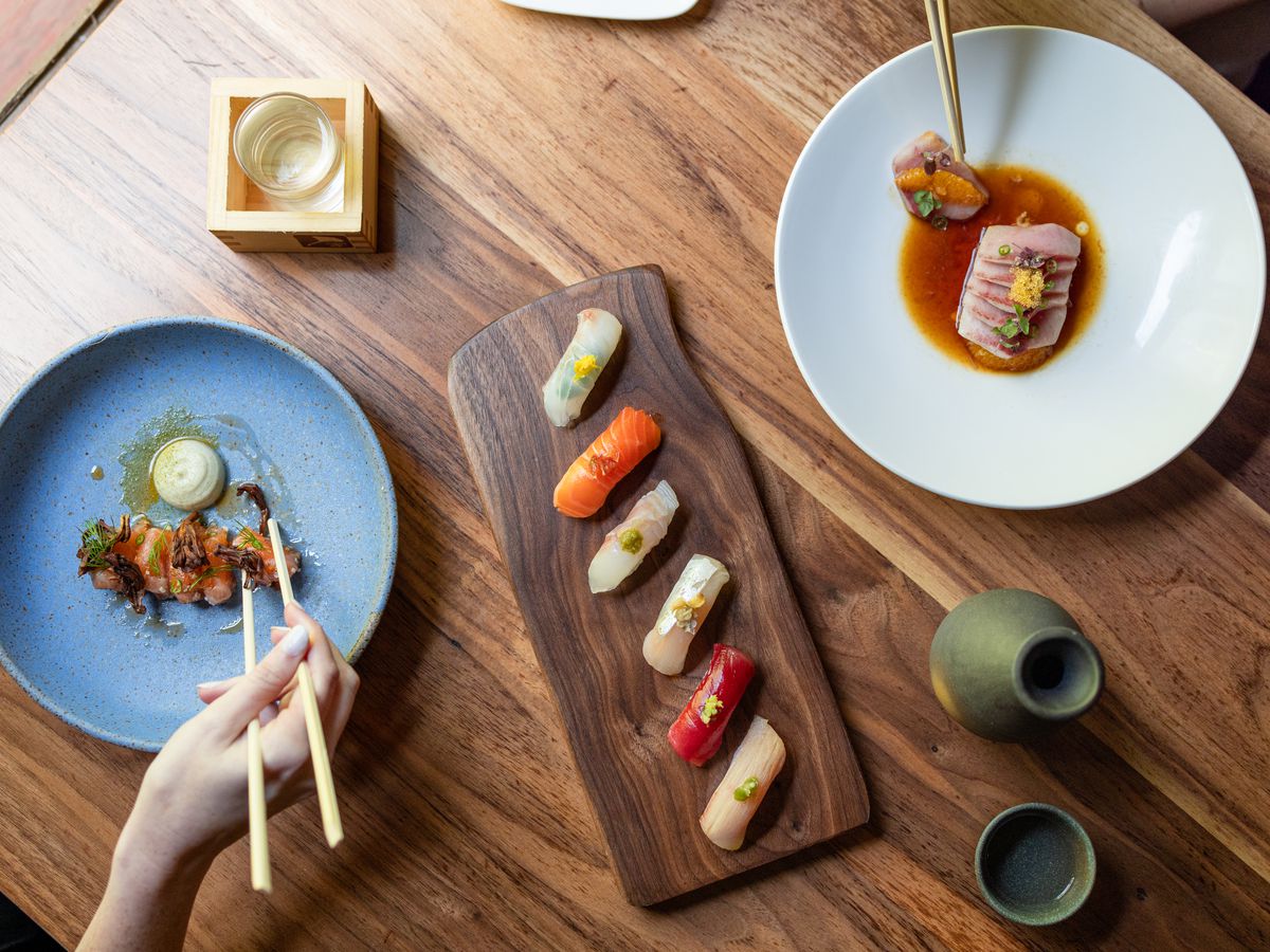 A wooden table holds (left to right) a blue plate with salmon on a creamy dip, a board with nigiri made with various fish, and a white plate with tuna in a dark sauce.