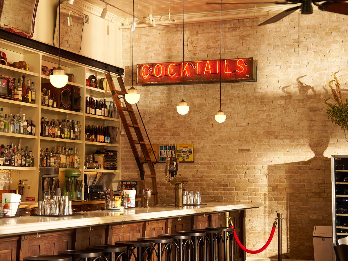 The interior of a bar has exposed brick walls and a bar back, with a neon sign reading “Cocktails” in red.