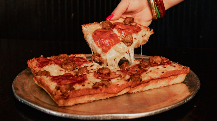 A hand lifts a slice of Detroit-style pizza from a pie sitting on a table.