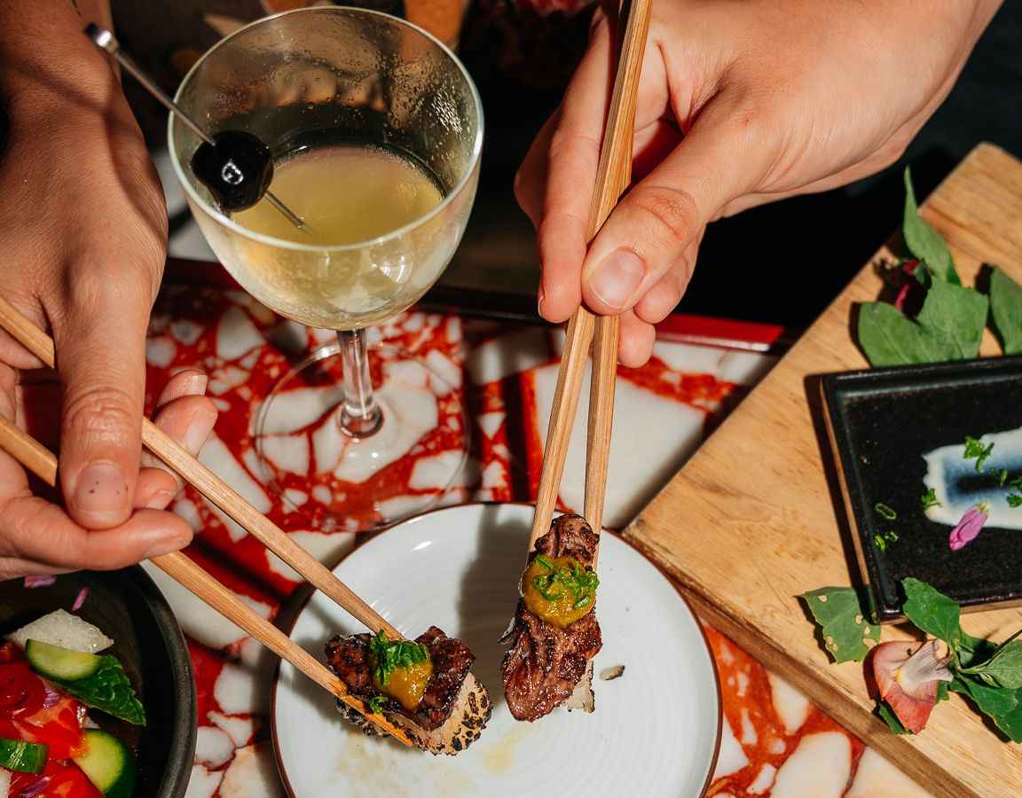 Two hands holding chopsticks reach for bites of food. A martini-like cocktail sits on the table above it.