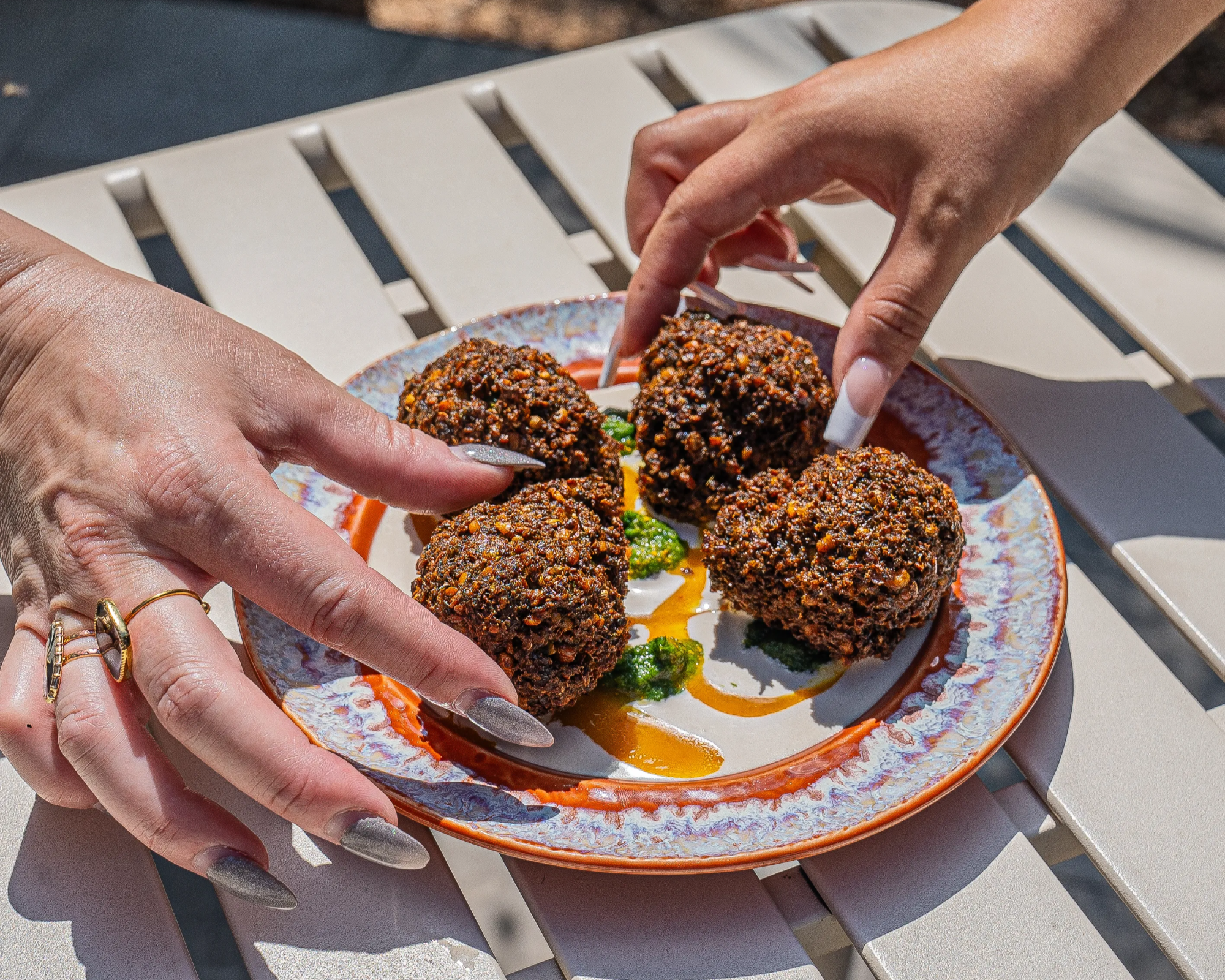 A plate holds four pieces of falafel. The hands of two women reach in to grab one each.